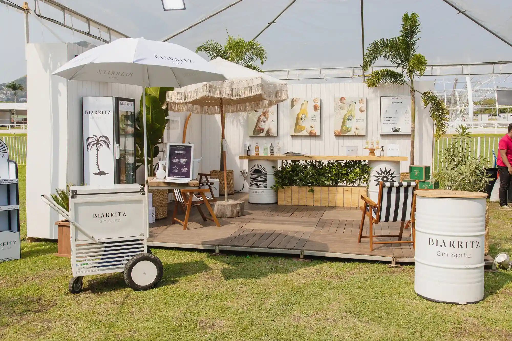 Um refrigerador branco com o rótulo “Biarritz Gin Spritz” em um balcão contém bebidas engarrafadas; um cardápio emoldurado e um refrigerador maior estão ao fundo.